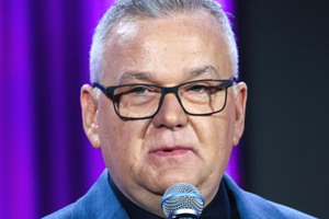 Artur Andrus mówiący do mikrofonu. Tło w kolorze fioletowym.