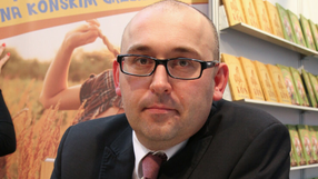 Mężczyzna w okularach z poważnym wyrazem twarzy, w garniturze, widoczny za stołem z książkami.