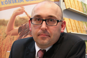 Mężczyzna w okularach z poważnym wyrazem twarzy, w garniturze, widoczny za stołem z książkami.