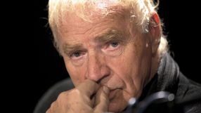 Osoba z jasnymi włosami, głęboko zamyślona, z ręką przy twarzy, w mrocznym tle.
