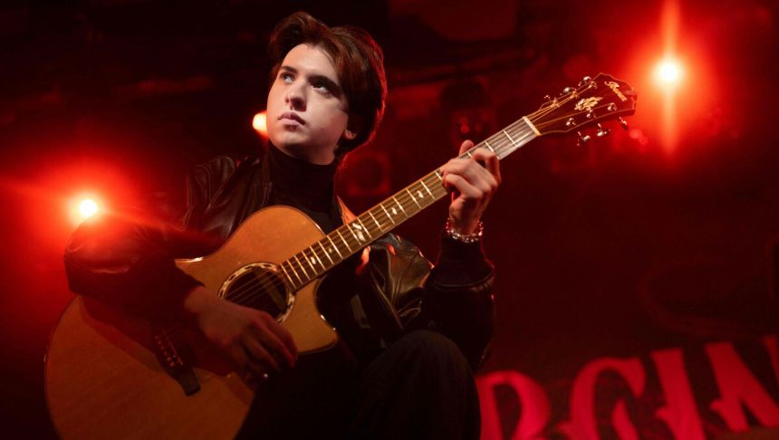 Młody mężczyzna gra na gitarze akustycznej, w tle czerwone światła sceny.