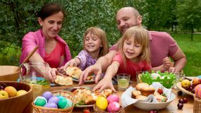 Rodzina bawi się przy stole, sięgając po jedzenie i dekoracyjne pisanki w słonecznym otoczeniu.