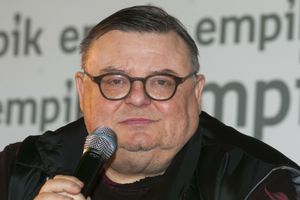 Mężczyzna trzyma mikrofon, ma okrągłe okulary i bladą cerę, siedzi na tle napisu Empik.