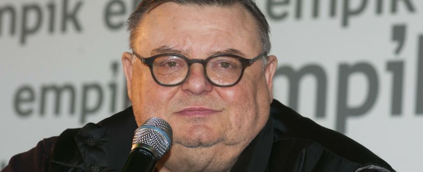 Mężczyzna trzyma mikrofon, ma okrągłe okulary i bladą cerę, siedzi na tle napisu Empik.