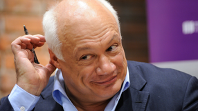 Mężczyzna z jasnymi włosami, uśmiechnięty, trzyma długopis przy uchu. W tle ceglana ściana.