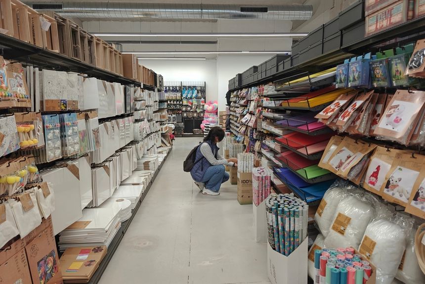 Osoba w dżinsach i kamizelce przegląda materiały w sklepie papierniczym między półkami.
