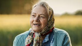 Starsza kobieta w chustce uśmiecha się na tle zielonego pola, promienie słońca oświetlają scenę.