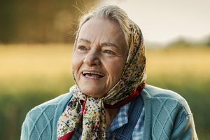 Starsza kobieta w chustce uśmiecha się na tle zielonego pola, promienie słońca oświetlają scenę.