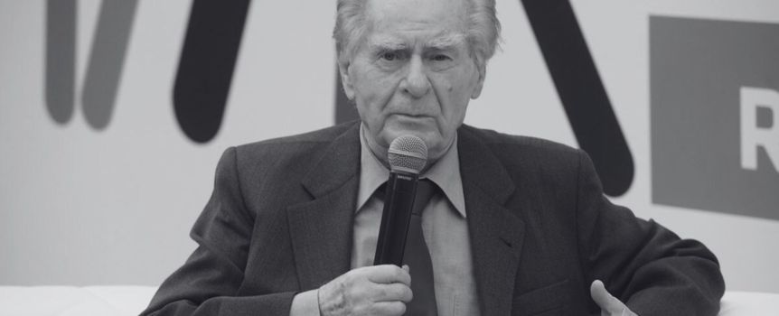Mężczyzna w szarej marynarce trzyma mikrofon, rozmawia na tle kolorowego tła.