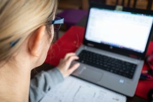 Osoba pracująca przy laptopie, zapisująca notatki na kartce, z widocznym biurkiem.