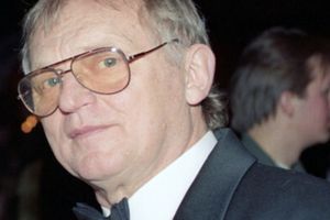 Mężczyzna w okularach i czarnym garniturze stoi na tle oświetlonego pomieszczenia.