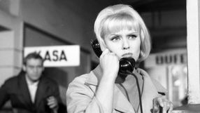Kobieta w jasnym płaszczu rozmawia przez czarną słuchawkę telefoniczną. 