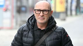 Mężczyzna w czarnej kurtce puchowej, z okularami, uśmiecha się na ulicy.