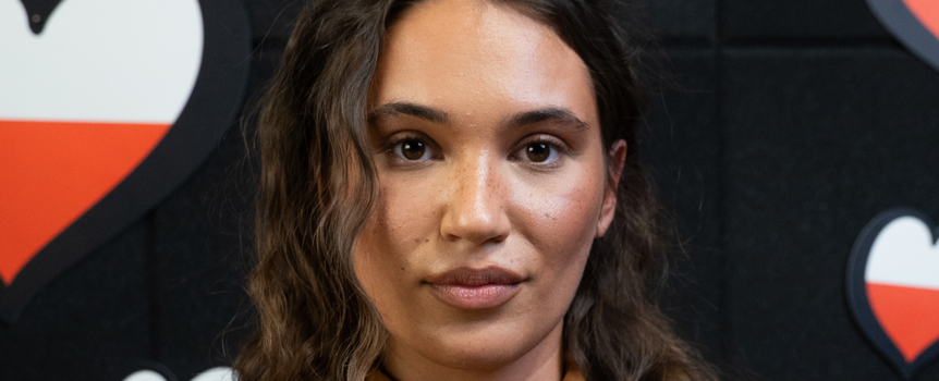 Kobieta z długimi kręconymi włosami, patrzy w obiektyw na tle serc w polskich barwach.