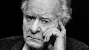 Wiesław Myśliwski. Mężczyzna z siwymi włosami, zamyślony, patrzy prosto w obiektyw, opierając rękę na twarzy.