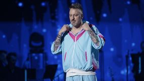 Piotr Kędzierski w kolorowym dresie, z tatuażami, na scenie wśród muzyków. Tło w nocnych kolorach.