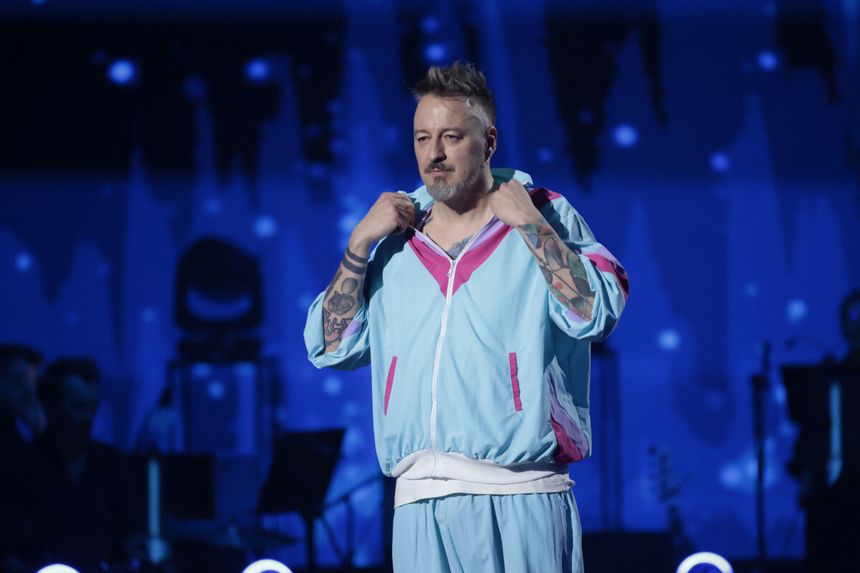 Piotr Kędzierski w kolorowym dresie, z tatuażami, na scenie wśród muzyków. Tło w nocnych kolorach.