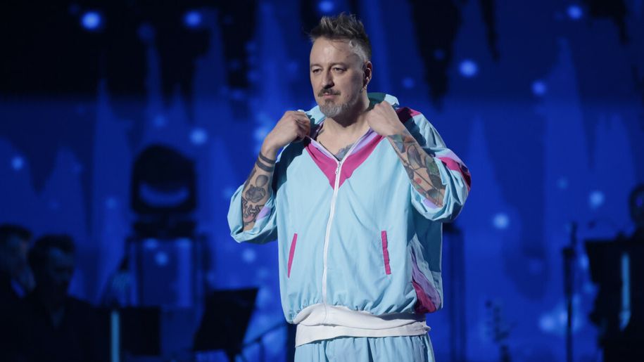 Piotr Kędzierski w kolorowym dresie, z tatuażami, na scenie wśród muzyków. Tło w nocnych kolorach.