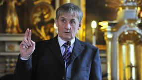Mężczyzna w garniturze mówi do mikrofonu, uniesiona ręka z palcami w geście powitania.
