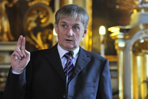 Mężczyzna w garniturze mówi do mikrofonu, uniesiona ręka z palcami w geście powitania.