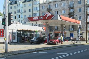 Stacja paliw Orlen z dwoma samochodami, w tle budynki mieszkalne i droga.