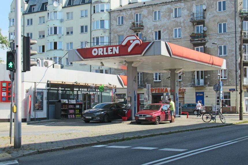 Stacja paliw Orlen z dwoma samochodami, w tle budynki mieszkalne i droga.