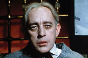 Mężczyzna z jasnymi włosami w swetrze, siedzi w ciemnym pomieszczeniu, patrząc w obiektyw.