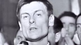 Mężczyzna mówi do mikrofonu, w tle widać zwolnione inne twarze, w atmosferze zgromadzenia.