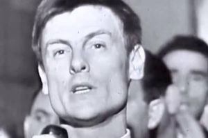 Mężczyzna mówi do mikrofonu, w tle widać zwolnione inne twarze, w atmosferze zgromadzenia.
