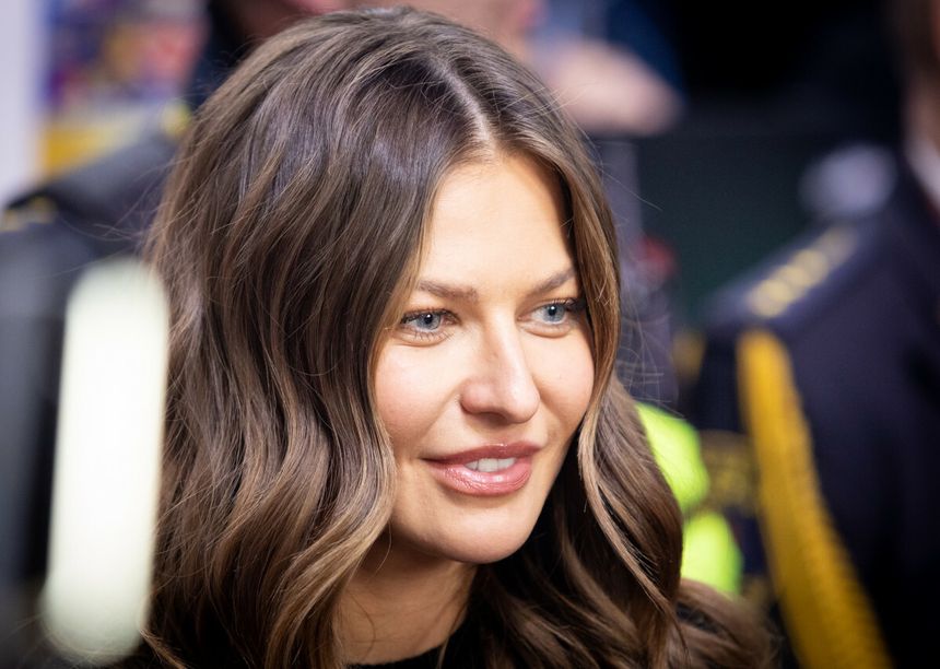 Na zdjęciu uśmiechająca się Anna Lewandowska z długimi, falowanymi włosami. W tle widoczni ludzie.