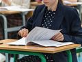 Uczennica w granatowym żakiecie przegląda dokumenty na egzaminie. W tle ławki szkolne.