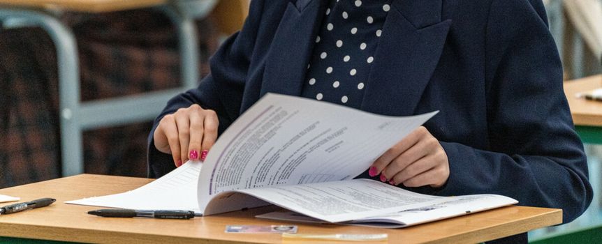 Uczennica w granatowym żakiecie przegląda dokumenty na egzaminie. W tle ławki szkolne.