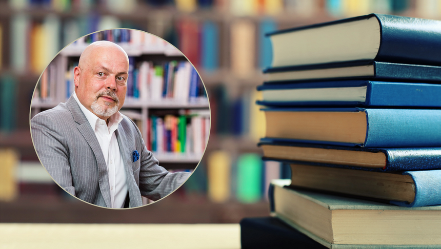Mężczyzna w jasnym garniturze, siedzi przed półką z książkami. W tle widoczne sterty książek.