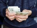 Mężczyzna w swetrze trzyma w rękach pakiet banknotów o nominale 100 zł.