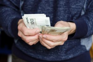 Mężczyzna w swetrze trzyma w rękach pakiet banknotów o nominale 100 zł.