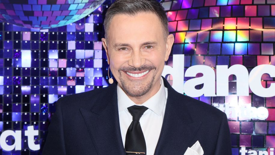Krzysztof Ibisz w garniturze, uśmiechnięty, na tle kolorowych dyskotekowych luster.