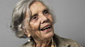 Starsza kobieta z uśmiechem, miękkimi, siwymi włosami, w perłowych kolczykach, na szarym tle.