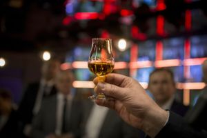 Szklanka z napojem trzymana w dłoni, w tle zamazane postacie ludzi i świetlne akcenty.