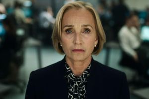 Kobieta z blond włosami i eleganckim strojem, patrząca w obiektyw, w tle biurowe wnętrze.