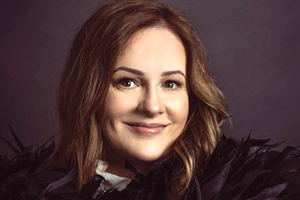 Kobieta z długimi, falującymi włosami, uśmiechająca się w eleganckim, puchatym swetrze.