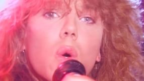 Mężczyzna z długimi, kręconymi włosami śpiewa do mikrofonu w różowym świetle.