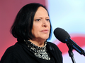 Grażyna Łobaszewska z ciemnymi włosami w czarnym swetrze, mówi do mikrofonu na tle jasnego tła.
