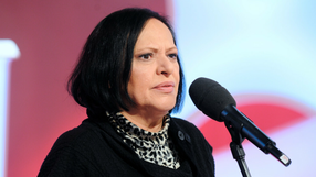 Grażyna Łobaszewska z ciemnymi włosami w czarnym swetrze, mówi do mikrofonu na tle jasnego tła.