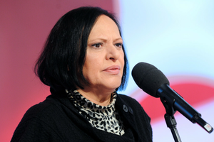 Grażyna Łobaszewska z ciemnymi włosami w czarnym swetrze, mówi do mikrofonu na tle jasnego tła.
