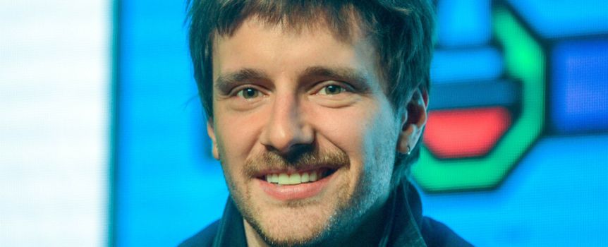 Uśmiechnięty mężczyzna z krótkimi włosami, w ciemnej kurtce, na tle kolorowego tła.