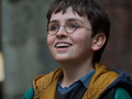 Harry Potter: Kulisy magii. Uśmiechnięty chłopak z okularami, w kolorowej kurtce, stoi na tle budynków.