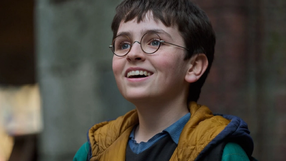Harry Potter: Kulisy magii. Uśmiechnięty chłopak z okularami, w kolorowej kurtce, stoi na tle budynków.