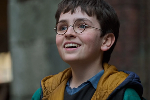 Harry Potter: Kulisy magii. Uśmiechnięty chłopak z okularami, w kolorowej kurtce, stoi na tle budynków.