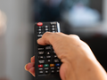 Na zdjęciu widać rękę trzymającą pilot telewizyjny, wskazującą na ekran telewizora.