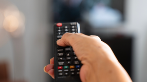 Na zdjęciu widać rękę trzymającą pilot telewizyjny, wskazującą na ekran telewizora.